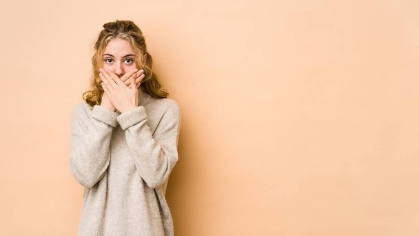 Young caucasian woman isolated on beige background shocked covering mouth with hands. - Foto, Imagem