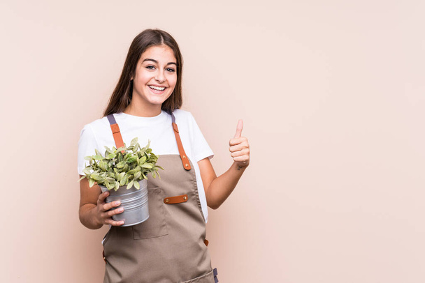 Young gardener caucasian woman holding a plant isolatedsmiling and raising thumb up - Фото, изображение