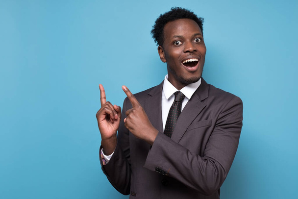 Zabawny młody, afrykański Amerykanin w garniturze, wskazujący palcem wskazującym w górę. Zwróć uwagę na tę koncepcję informacji. Studio nakręcone na niebieskiej ścianie. - Zdjęcie, obraz