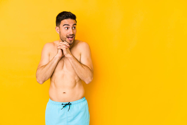 Junger kaukasischer Mann mit Badeanzug isoliert auf gelbem Hintergrund Junger kaukasischer Mann mit trinkendem Glück, erstaunt und öffnendem Mund, der nach vorne schaut. - Foto, Bild