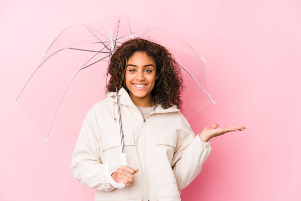 Joven mujer afroamericana sosteniendo un paraguas mostrando un espacio de copia en una palma y sosteniendo otra mano en la cintura. - Foto, Imagen