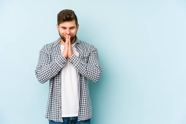Jonge blanke man geïsoleerd op blauwe achtergrond hand in hand bidden in de buurt van mond, voelt vertrouwen. - Foto, afbeelding