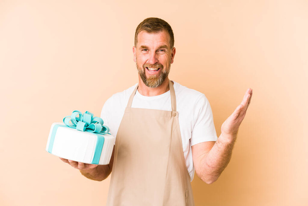 Senior sosteniendo un pastel aislado sobre fondo beige recibiendo una agradable sorpresa, excitado y levantando las manos. - Foto, imagen