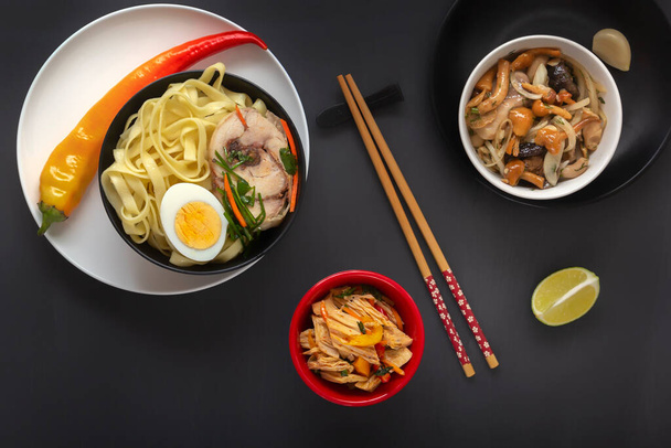 Noodles with pieces of meat and egg in a black plate and dim sums with different traditional snacks of mushrooms, soy and vegetables with chopsticks on a dark table. View from above. Asian food concept - Foto, Imagen