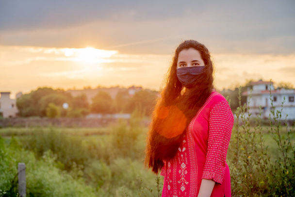 Giovane ragazza indiana indossando una maschera, indiano salwar kameez in piedi con un campo e un bel tramonto dietro di lei - Foto, immagini