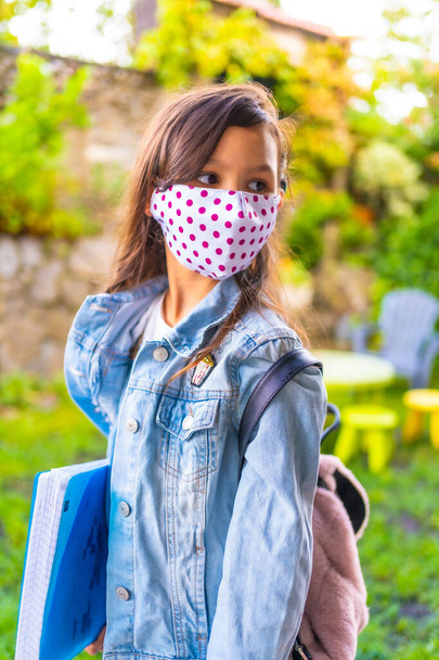 Caucasian girl with face mask ready to go back to school. New normality, social distance, coronavirus pandemic, covid-19. Jacket, backpack, mask with pink dots and a blue block in hand - Foto, Imagem