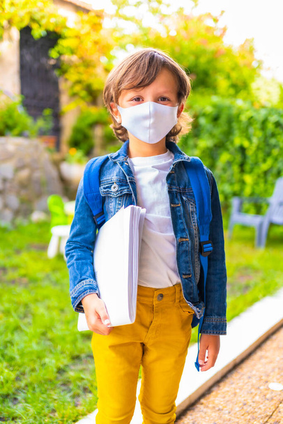 Caucasian child with face mask ready for back to school. New normality, social distance, coronavirus pandemic, covid-19. With denim jacket, backpack and notepad in hand - Foto, immagini