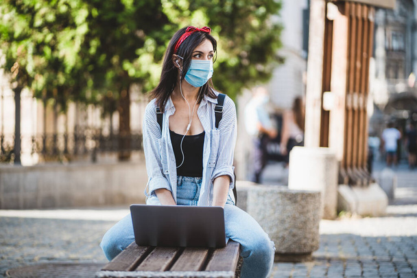 Εστιασμένη νεαρή γυναίκα κοιτάζει πλάγια, φοράει προστατευτική μάσκα προσώπου, κάθεται σε δημόσιο παγκάκι και χρησιμοποιεί φορητό υπολογιστή. Εκπαίδευση, Απομακρυσμένη Εργασία, Πανδημία και Νέα Κανονική Έννοια - Φωτογραφία, εικόνα