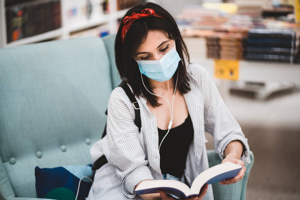 Egy fiatal nő védőmaszkot visel és könyvet olvas, miközben a könyvtárban vagy könyvesboltban ül a kanapén. Életmód, oktatás és új normális koncepció a koronavírus világjárvány idején - Fotó, kép