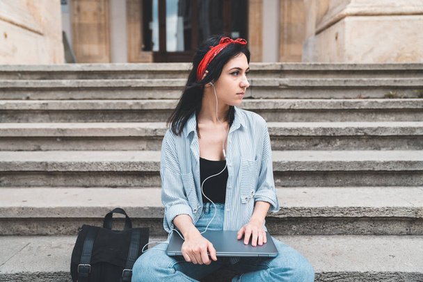 Młoda kobieta siedząca na schodach, patrząca bokiem, trzymająca laptopa i słuchająca muzyki ze słuchawek w kampusie uniwersyteckim. Koncepcja stylu życia, edukacji i pandemii - Zdjęcie, obraz