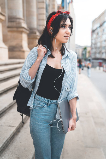 Молода стильна жінка ходить і оглядає університетське містечко, тримаючи ноутбук і слухаючи музику навушників. Красива жінка в блакитній сорочці і джинсах. Концепція способу життя та освіти
. - Фото, зображення
