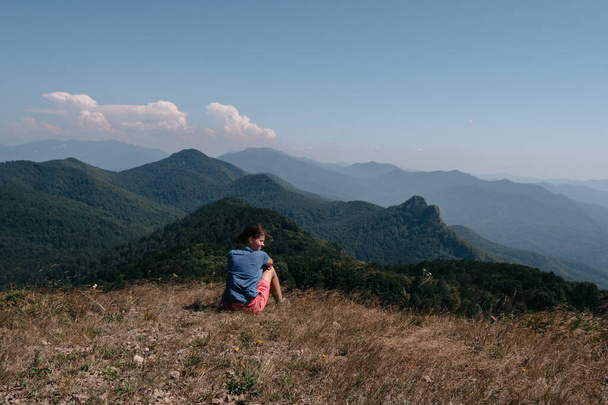 Μια γυναίκα ταξιδιώτης κάθεται στην κορυφή ενός λόφου και κοιτάζει τα γύρω βουνά. Όμορφο ορεινό και λοφώδες τοπίο σε ζεστό, ηλιόλουστο καιρό και ο νεαρός τουρίστας. - Φωτογραφία, εικόνα