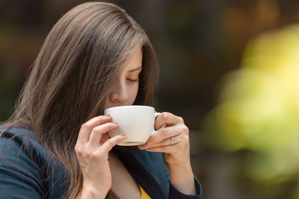 young white-skinned woman with long, straight hair and makeup, keeps her eyes closed while taking a white cup with both hands, wearing a ring and a blue jacket, the background is blurred - Foto, Imagem