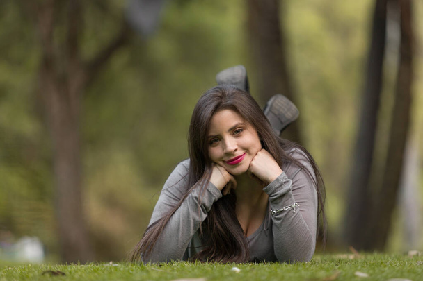 joven mujer de piel blanca con el pelo largo y maquillaje se encuentra boca abajo en la hierba con las manos en la barbilla, sonriendo y usando una pulsera, blusa gris en un parque - Foto, Imagen