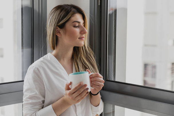 Young beautiful woman near window with cup of coffee or tea at home. Dream and relax. Good day. Woman in the morning is holding a mug and looking at the sunrise and having a perfect cozy morning - Fotografie, Obrázek