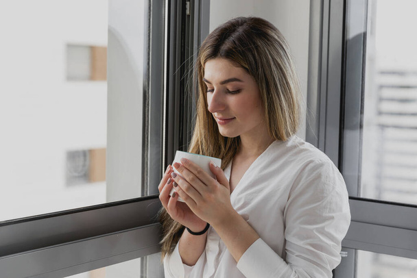 Jovem mulher bonita perto da janela com xícara de café ou chá em casa. Sonhe e relaxe. Bom dia. Mulher de manhã está segurando uma caneca e olhando para o nascer do sol e ter uma manhã aconchegante perfeito - Foto, Imagem