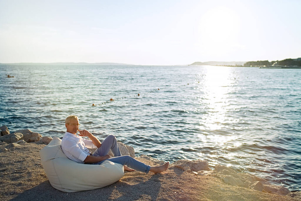 Senior businessman sitting on deck chair on the beach during sunset - Foto, imagen