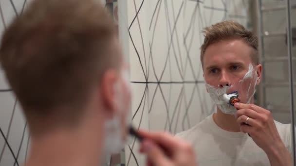 A blond man shaves his stubble after applying shaving foam in a white bathroom - Footage, Video