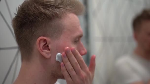 Un hombre rubio se afeita la barba después de aplicar espuma de afeitar en un baño blanco - Metraje, vídeo