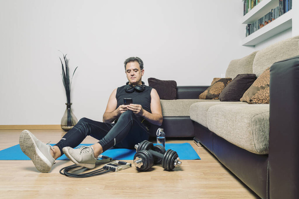 обычный зрелый мужчина собирается заниматься спортом дома, используя приложение для смартфонов. Упражнения в одиночку в квартире. Старайтесь оставаться здоровым и здоровым с хорошим телом - Фото, изображение
