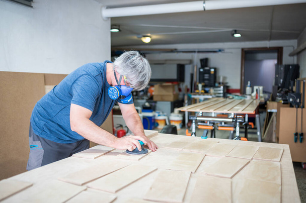 Carpinteiro maduro usando máscara lixadeira enquanto trabalhava em peças de madeira.Copiar espaço - Foto, Imagem