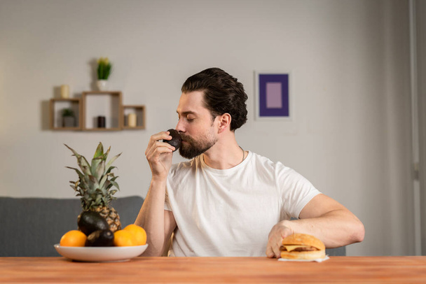 Een jonge man zit aan een tafel en praat over het nut van fruit, hij pakt ze op en toont avocado. Gezond voedsel - Foto, afbeelding