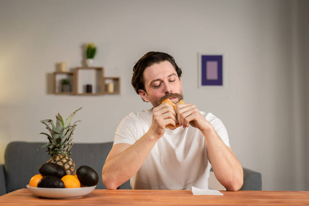 Un jeune homme s'assied à une table et parle de nourriture, il choisit le hamburger et le mange - Photo, image