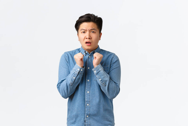 Portrait of timid and insecure young asian man feeling cornered or scared, holding hands tight to chest, shivering fear, looking anxious at camera, standing frightened over white background - Photo, Image