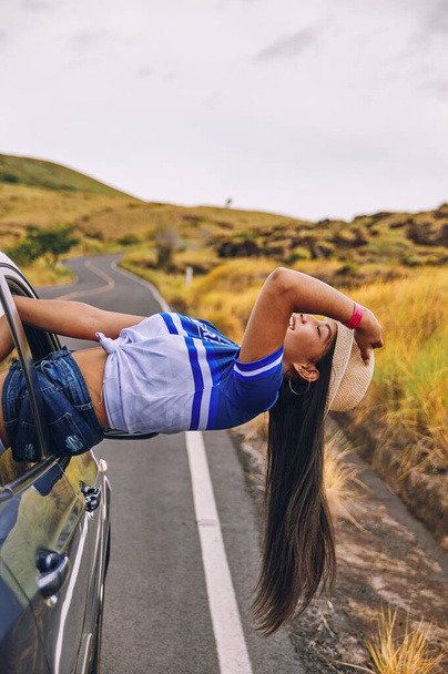 Eine fröhliche junge Frau mit Hut sitzt lächelnd im Fenster der Autotür und genießt die Fahrt und hat Spaß - Foto, Bild