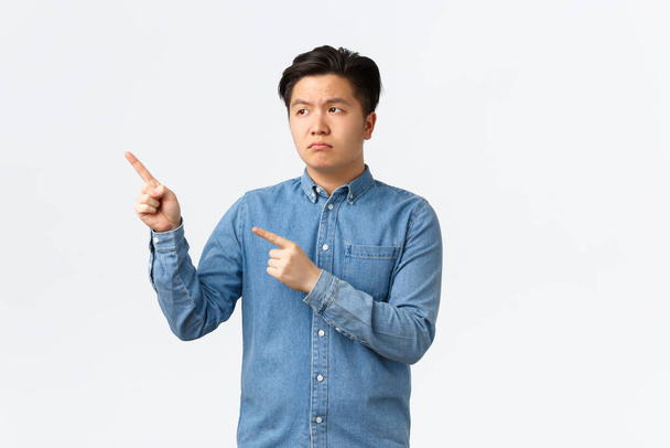 Unamused und enttäuscht asiatischer düsterer Mann in blauem Hemd, runzelt die Stirn und zeigt mit dem Finger in die linke obere Ecke, beschwert sich über schlechtes Produkt, fühlt sich bedrängt, steht weißer Hintergrund - Foto, Bild