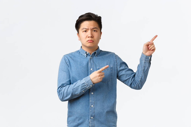 疑わしいと疑わしいアジアの男性が青いシャツを着て、懐疑的で不満を言い、指を右上隅に向け、質問をし、何か問題があると不平を言う、白い背景 - 写真・画像