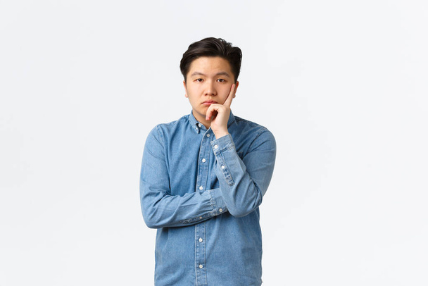 Znudzony i niezainteresowany azjatycki mężczyzna w niebieskiej koszuli, wyglądający niewesoło i niedbale przed kamerą, słuchający nudnej mowy, stojący niezadowolony i zirytowany białym tłem, zmęczony osobą - Zdjęcie, obraz