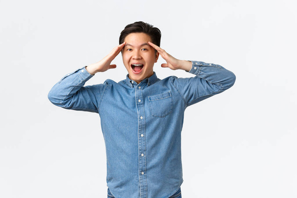 Surprised and happy, relieved asian man rejoicing over great news, holding hands near head and smiling upbeat, got rid of problem, feeling cheerful and upbeat over white background - Photo, Image