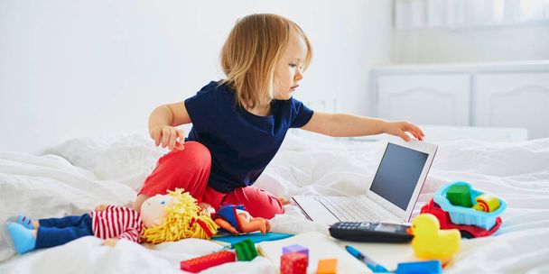 Toddler girl with laptop, notebook, phone and different toys in bed on clean white linens. Freelance, distance learning or work from home with kids concept - Fotoğraf, Görsel