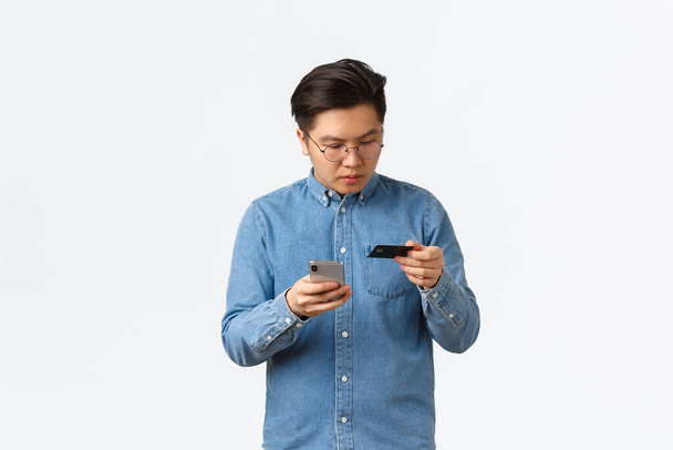 Koncentrált ázsiai fickó szemüvegben beírja a hitelkártya számát, hogy online vásárlás, a mobiltelefon és a bankszámla vásárolni valamit az interneten áruház, álló fehér háttér - Fotó, kép
