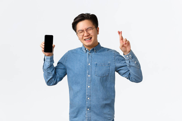 Hoffnungsvoller asiatischer Kerl mit Zahnspange, geschlossenen Augen und Daumen viel Glück, zeigt Smartphone-Bildschirm, wartet auf Antwort auf Dating-App, bettelt Gott an, steht vor weißem Hintergrund und macht Wünsche - Foto, Bild