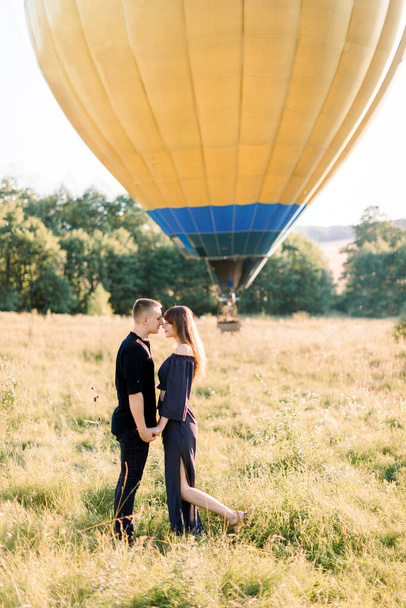 Couple in love stands face to face, holding hands, in summer field on the background of yellow air balloon. Man and woman in black clothes, celebrating their anniversary with flying on balloon - Foto, immagini