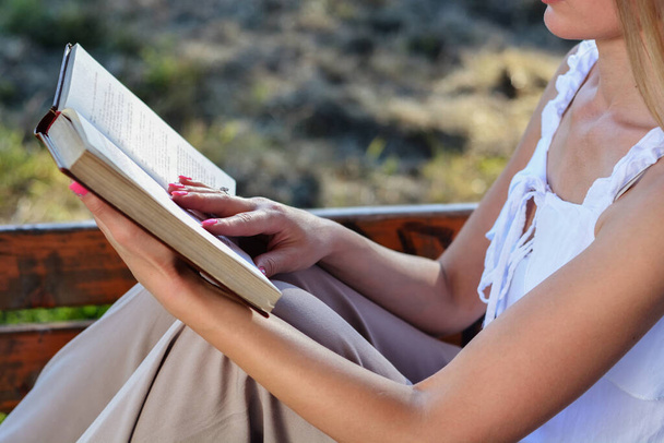 Frau hält ein aufgeschlagenes Buch in der Hand und liest. Nahaufnahme eines Mädchens, das an einem Sommertag auf einer Bank im Park sitzt - Foto, Bild