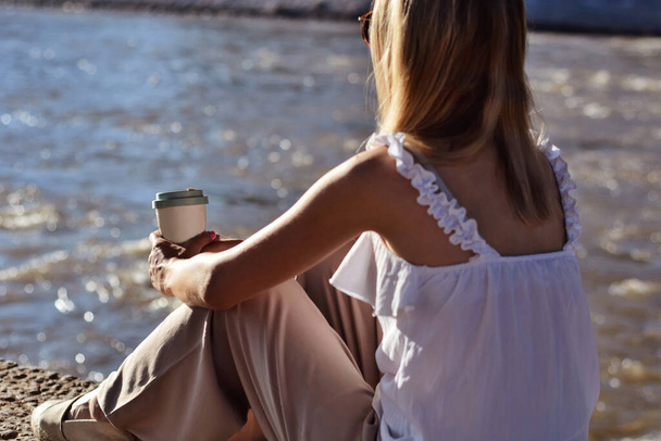 Mulher segurando uma xícara de café na mão e sentada ao lado do rio em um dia ensolarado de verão ao pôr do sol. Relaxamento no conceito de natureza - Foto, Imagem