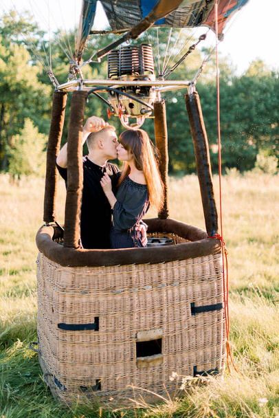 Young beautiful couple in black clothes, kissing in the basket of hot air balloon, enjoying their first fly in warm summer sunrise in the field - Foto, Imagem