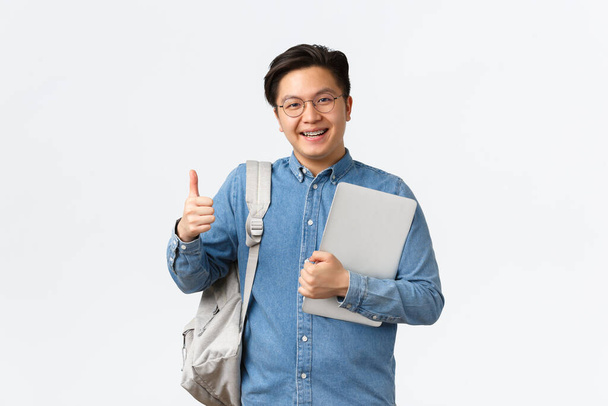 Universidad, estudiar en el extranjero y el concepto de estilo de vida. Satisfecho feliz asiático macho estudiante en gafas y camisa mostrando los pulgares hacia arriba en la aprobación, le gusta estudiar en la universidad, sosteniendo portátil y mochila - Foto, imagen
