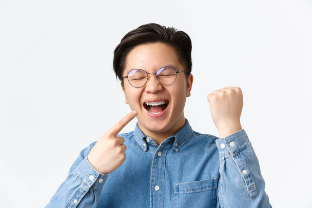 Ortodonzia e concetto di stomatologia. Primo piano di soddisfatto felice uomo asiatico che punta al suo apparecchio dentale e sorridente ampiamente, pompa pugno, gioire, fissaggio dei denti, in piedi sfondo bianco - Foto, immagini