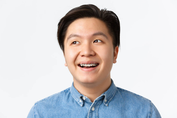 Orthodontics, dental care and stomatology concept. Close-up of hopeful, handsome asian man with teeth braces looking dreamy upper left corner, smiling pleased, standing white background - Photo, Image