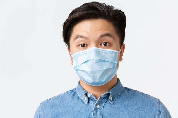 Coronavirus, soziale Distanzierung und Lebensstilkonzept. Überraschter asiatischer Mann in medizinischer Maske zieht ehrfurchtsvoll die Augenbrauen hoch, steht erstaunt über weißem Hintergrund und nutzt Schutzmaßnahmen während des Covid-19. - Foto, Bild