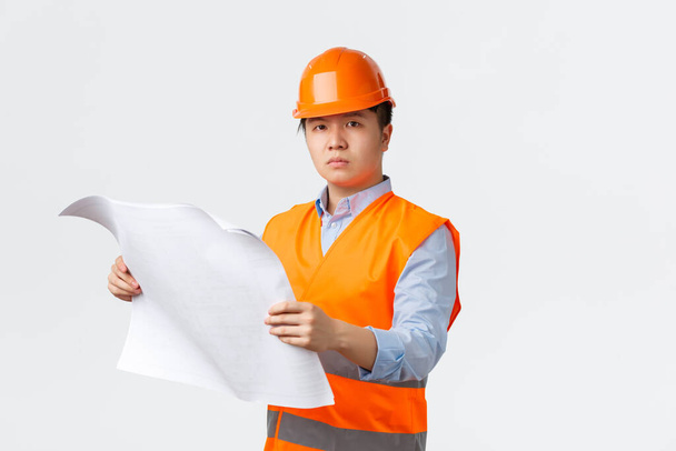 Building sector and industrial workers concept. Serious-looking confident asian construction manager, engineer in helmet and reflective jacket looking at blueprint, checking layout, white background - Foto, Imagen