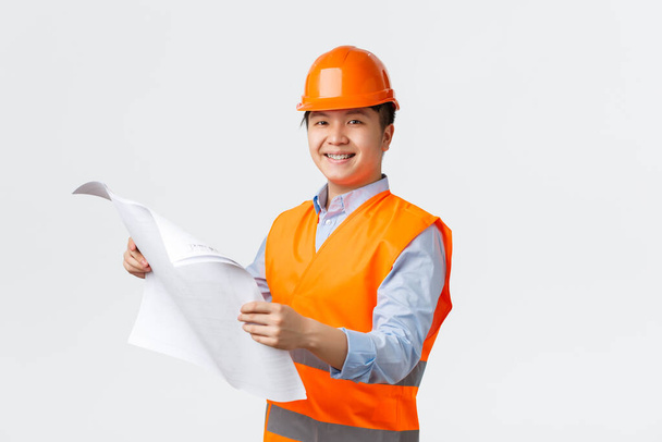 Building sector and industrial workers concept. Confident smiling asian architect, chief engineer in helmet and reflective jacket holding blueprints, inspecting enterprise, white background - Valokuva, kuva