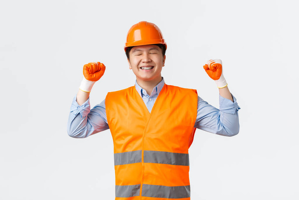 İnşaat sektörü ve endüstriyel işçiler kavramı. Başarılı Asyalı baş mühendis işini başarıyla tamamladı, mutlu görünüyor, kutlama için yumruk tokuşturuyor, gülümsüyor, miğfer ve güvenlik eldiveni takıyor. - Fotoğraf, Görsel