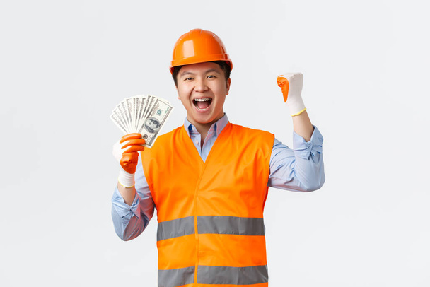 Koncept odvětví stavebnictví a pracovníků v průmyslu. Happy triumfující asijský stavební manažer, architekt v helmě a reflexní oblečení, pěst čerpadlo říká ano a ukazuje peníze, bílé pozadí - Fotografie, Obrázek