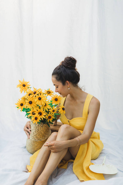 Portrait of young brunette woman on background of white hanging textile cloth with straw basket of yellow flowers, summer linen dress. - Photo, Image