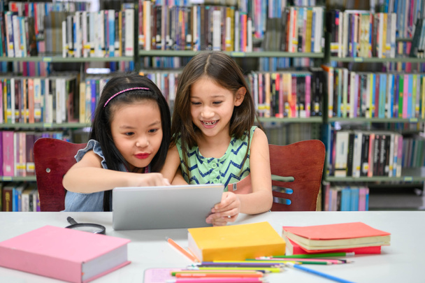 Δύο μικρά χαρούμενα χαριτωμένα κορίτσια που παίζουν σε μια συσκευή υπολογιστών tablet στη βιβλιοθήκη στο σχολείο. Εκπαίδευση και αυτο-μάθηση έννοια της ασύρματης τεχνολογίας. Ο τρόπος ζωής και η φιλία των ανθρώπων. Νηπιαγωγεία - Φωτογραφία, εικόνα